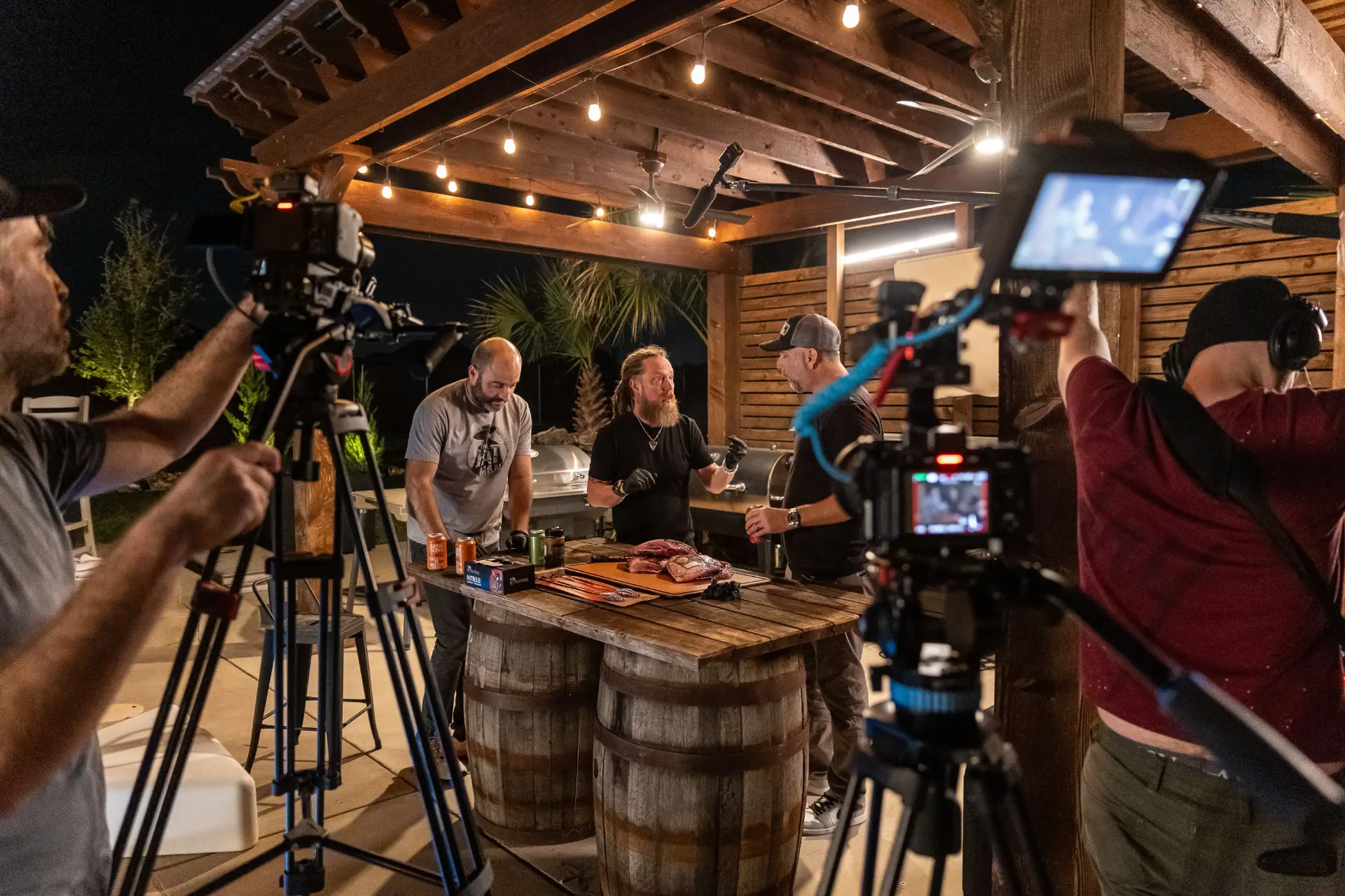 The hosts filming a night barbecue cook under string lights with camera crew