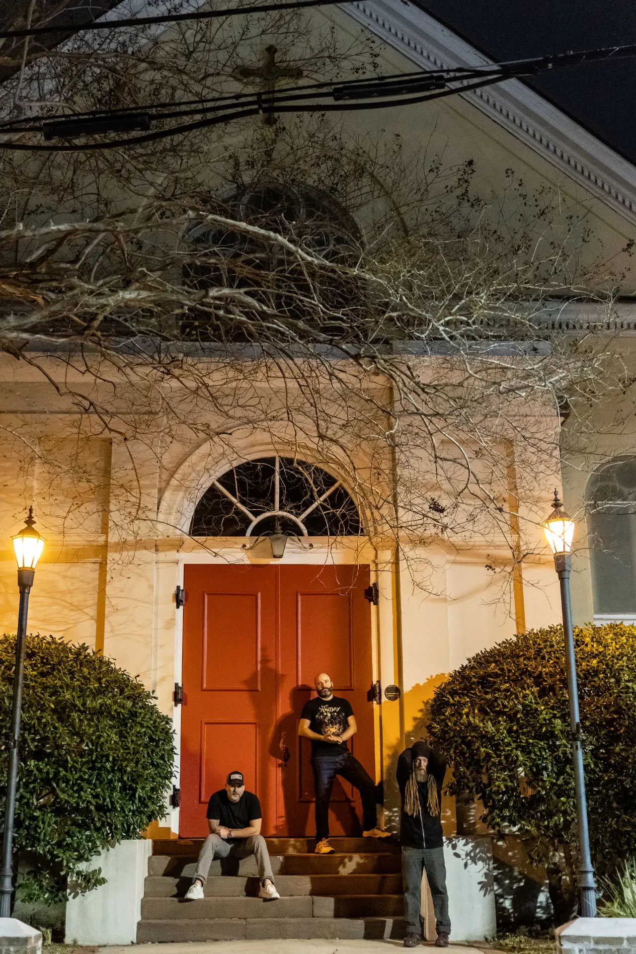The three hosts on the steps of a historic church at night beneath bare trees