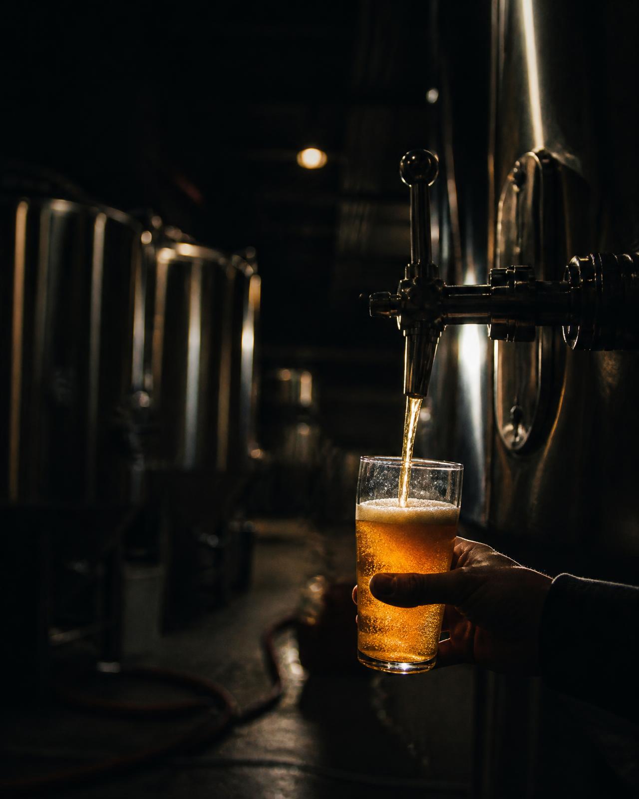 A glass of amber beer being poured in a dim brewery